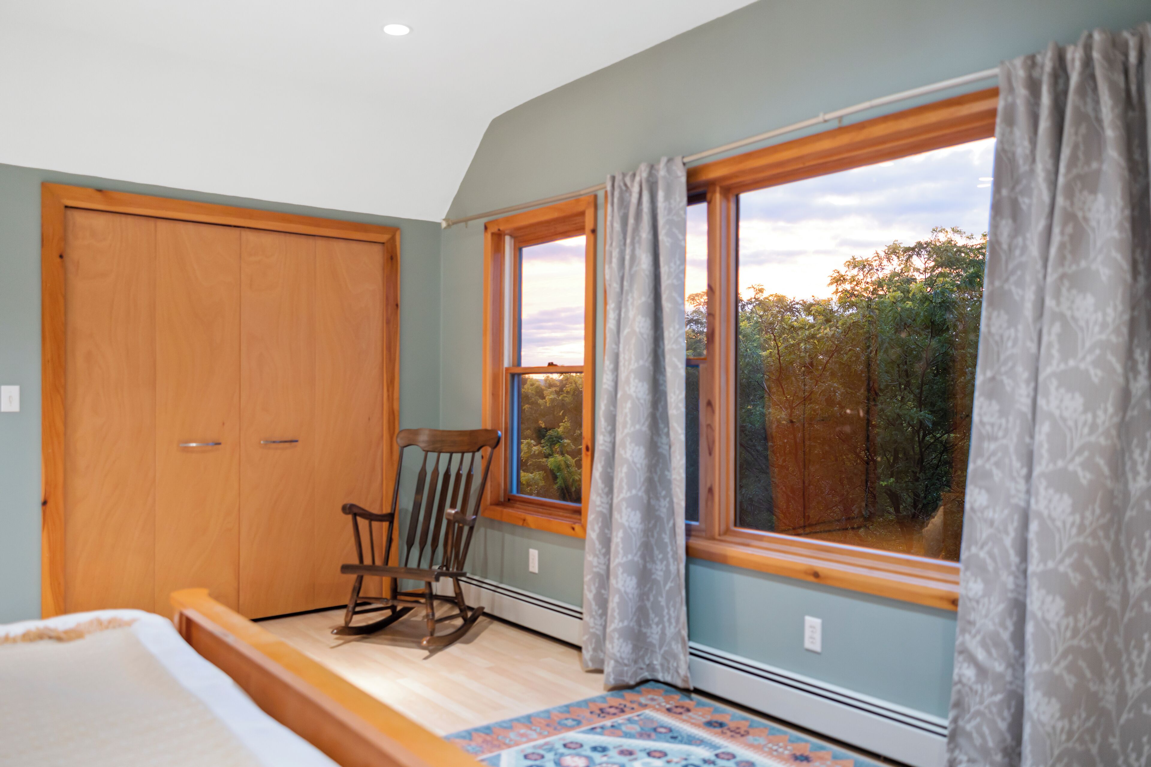 A bedroom with a wooden wardrobe, a rocking chair, a large window with curtains, and a patterned rug.