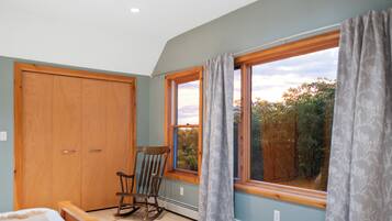 A bedroom with a wooden wardrobe, a rocking chair, a large window with curtains, and a patterned rug.