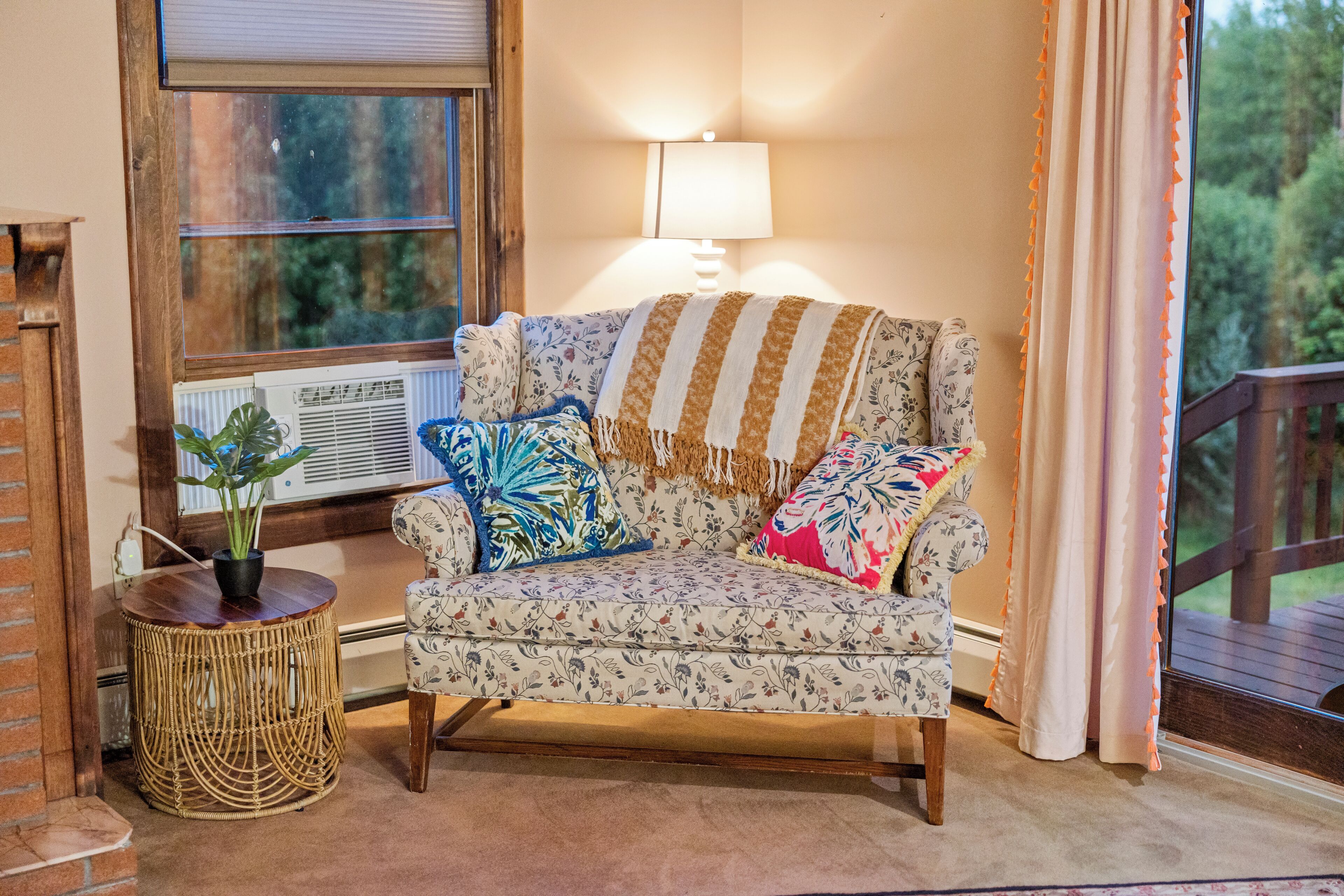 A cozy living room with a floral-patterned sofa, a round wicker side table with a potted plant, and a window with a view of greenery.