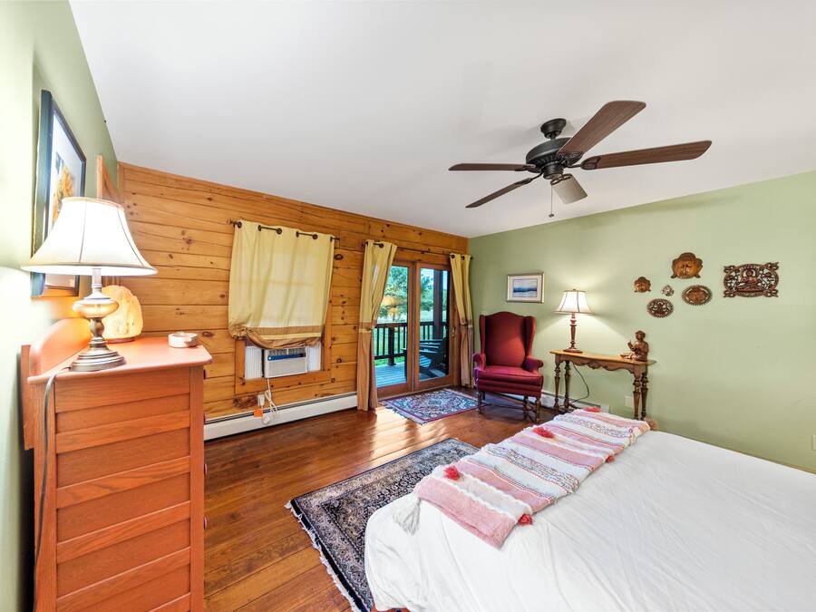 Quarto com paredes de madeira, ventoinha no teto, cama com manta estampada, escrivaninha com luminária e uma cadeira.