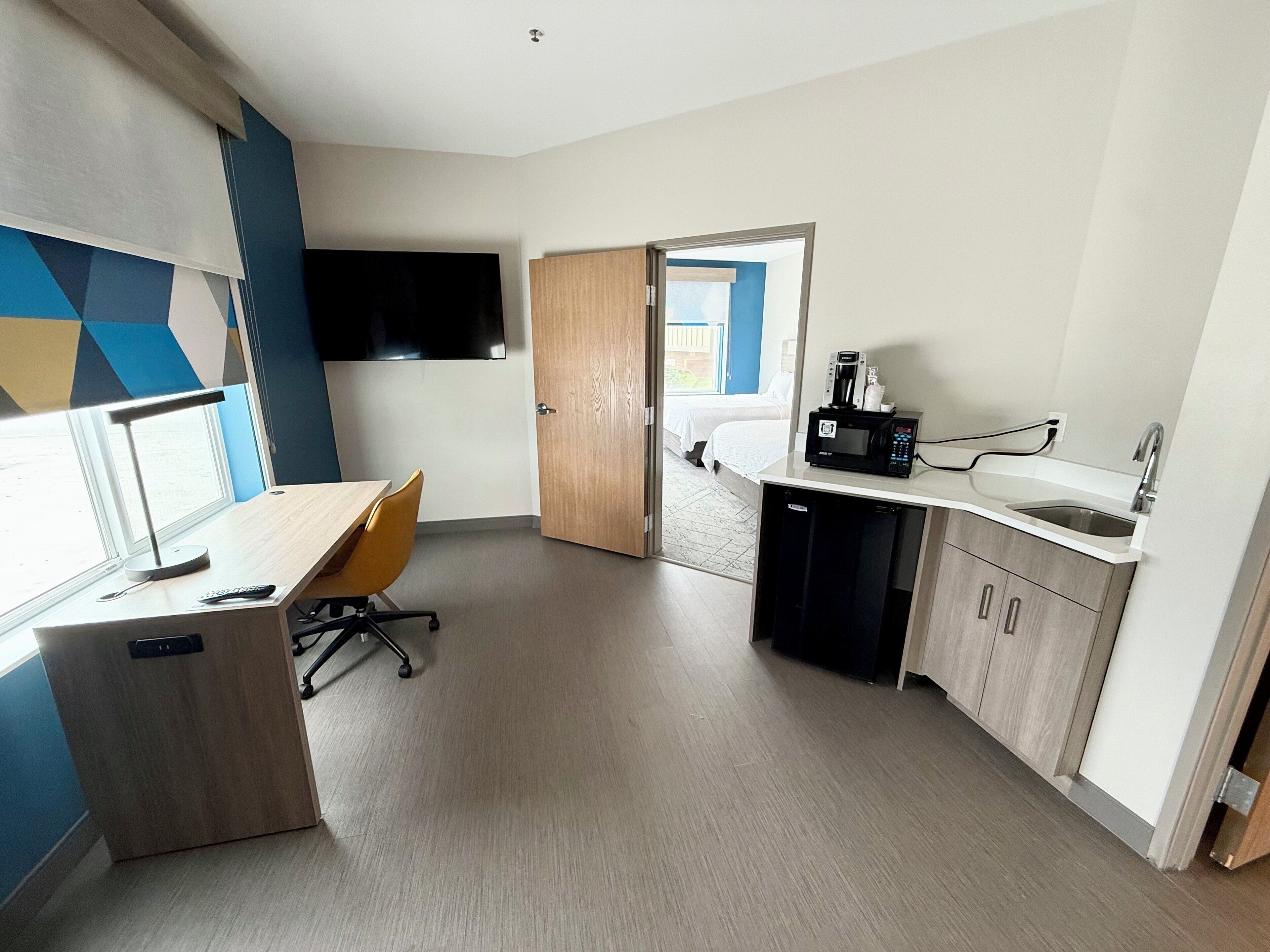 Ein modernes Hotelzimmer mit einem Holztisch, einem Stuhl, einem Flachbildfernseher, einer Mikrowelle und einem Waschbecken.