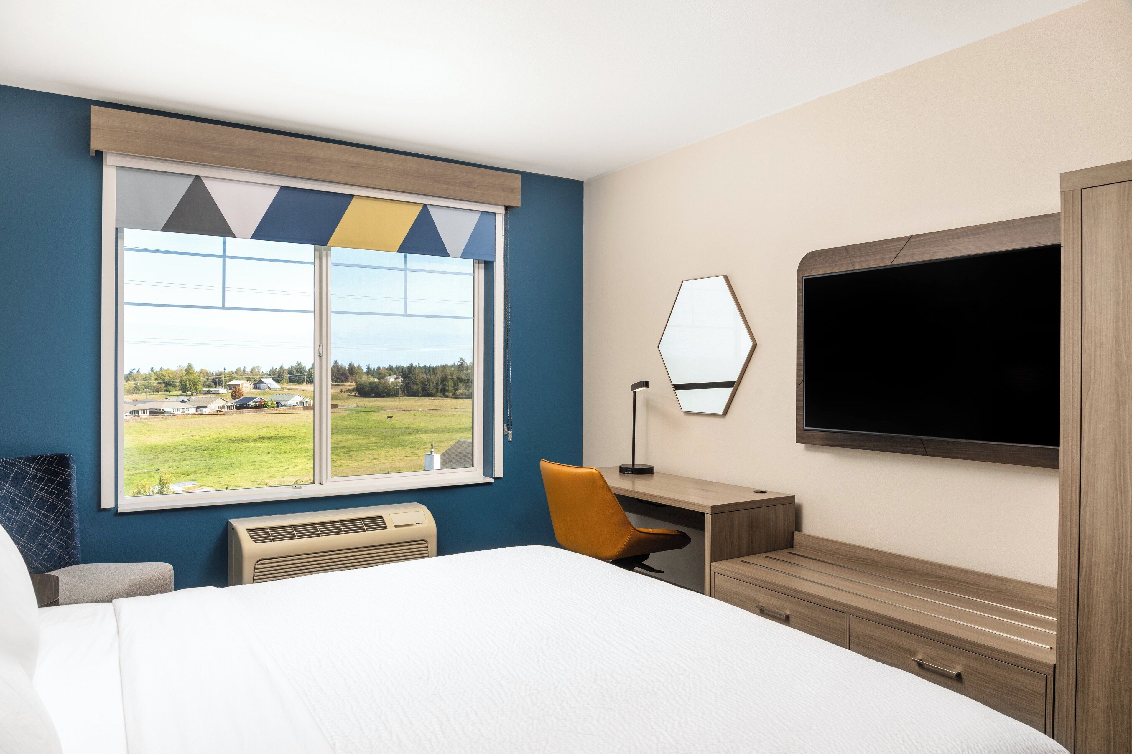 A hotel room with a large bed, a desk, a chair, a flat-screen TV, and a window with a view of a green landscape.