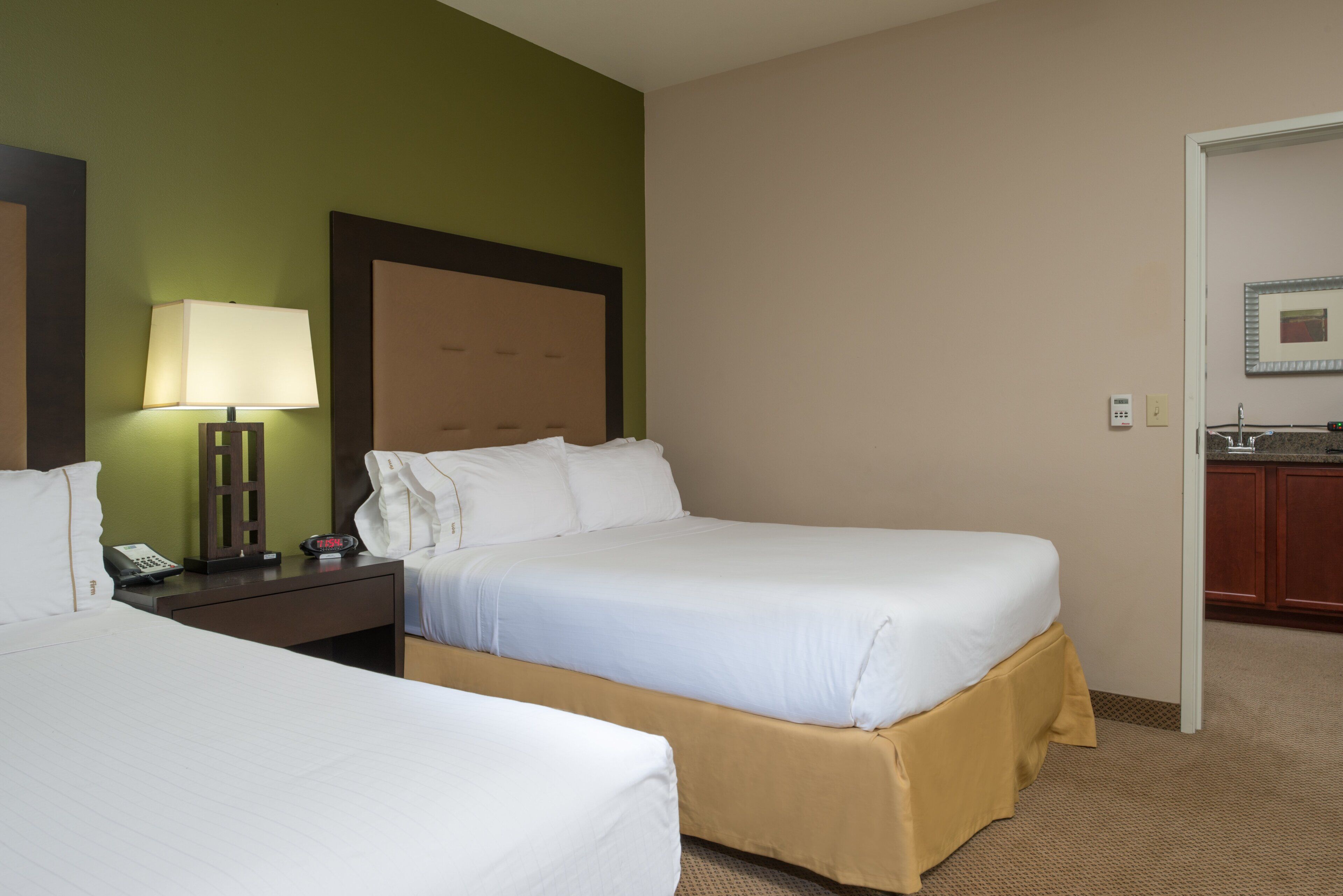 A hotel room with two beds, a nightstand with a lamp, a clock, and a small table.