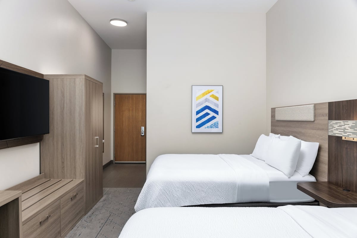 Ein Hotelzimmer mit zwei Betten, einem Fernseher, einem Kleiderschrank und einem gerahmten Kunstwerk an der Wand.
