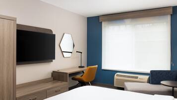 A hotel room with a bed, a desk, a chair, a television, a mirror, and a large window with blinds.