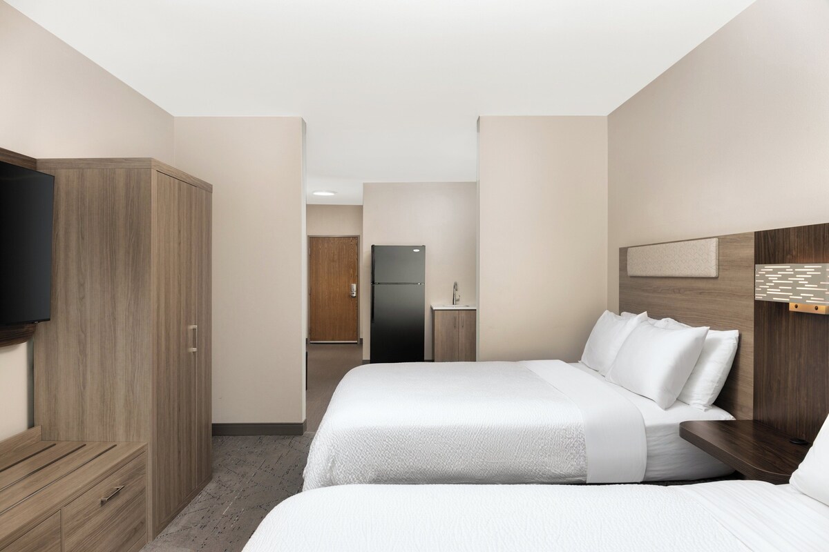 A hotel room with two beds, a wardrobe, a television, a small kitchenette, and a bathroom door.
