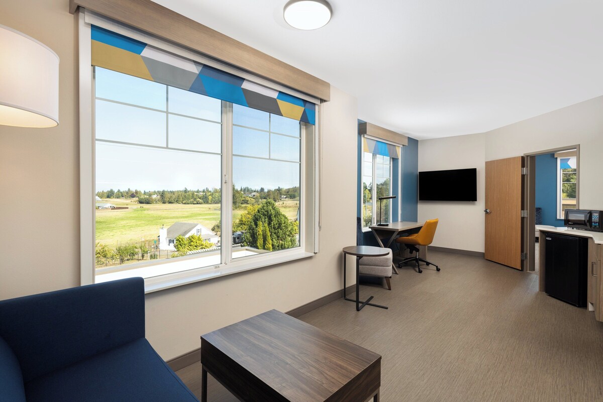 A modern hotel room with large windows, a flat-screen TV, a desk, and a comfortable seating area.