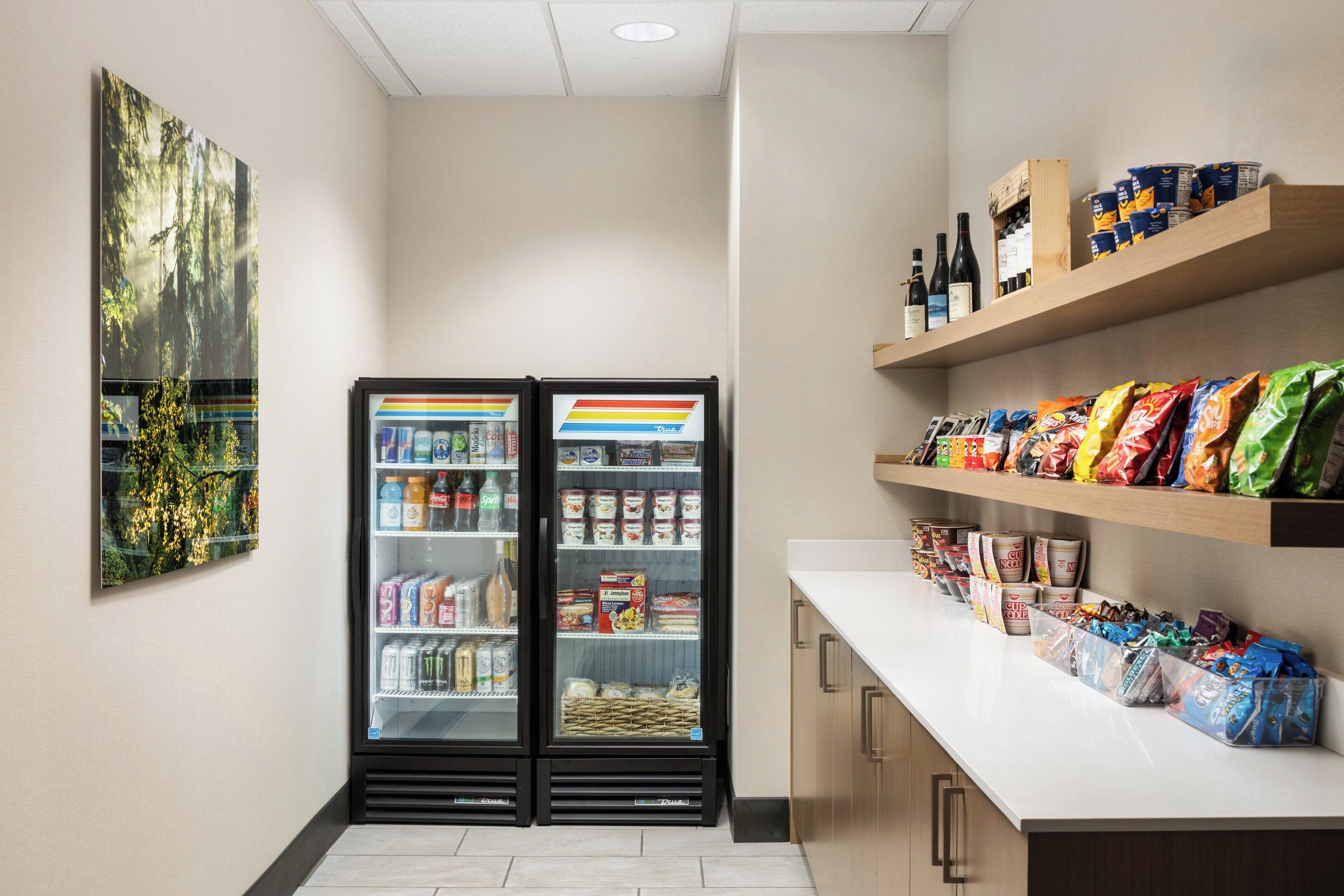 A clean, well-lit room with a refrigerator stocked with beverages, a snack bar with various packaged foods, and a wall-mounted painting.