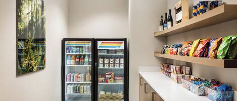 A clean, well-lit room with a refrigerator stocked with beverages, a snack bar with various packaged foods, and a wall-mounted painting.