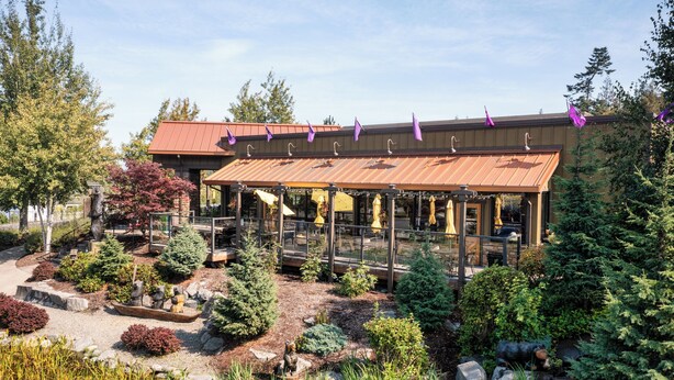 A building with a covered outdoor seating area, surrounded by landscaped gardens and trees. - Holiday Inn Express Hotel & Suites NORTH SEQUIM by IHG (Sequim)