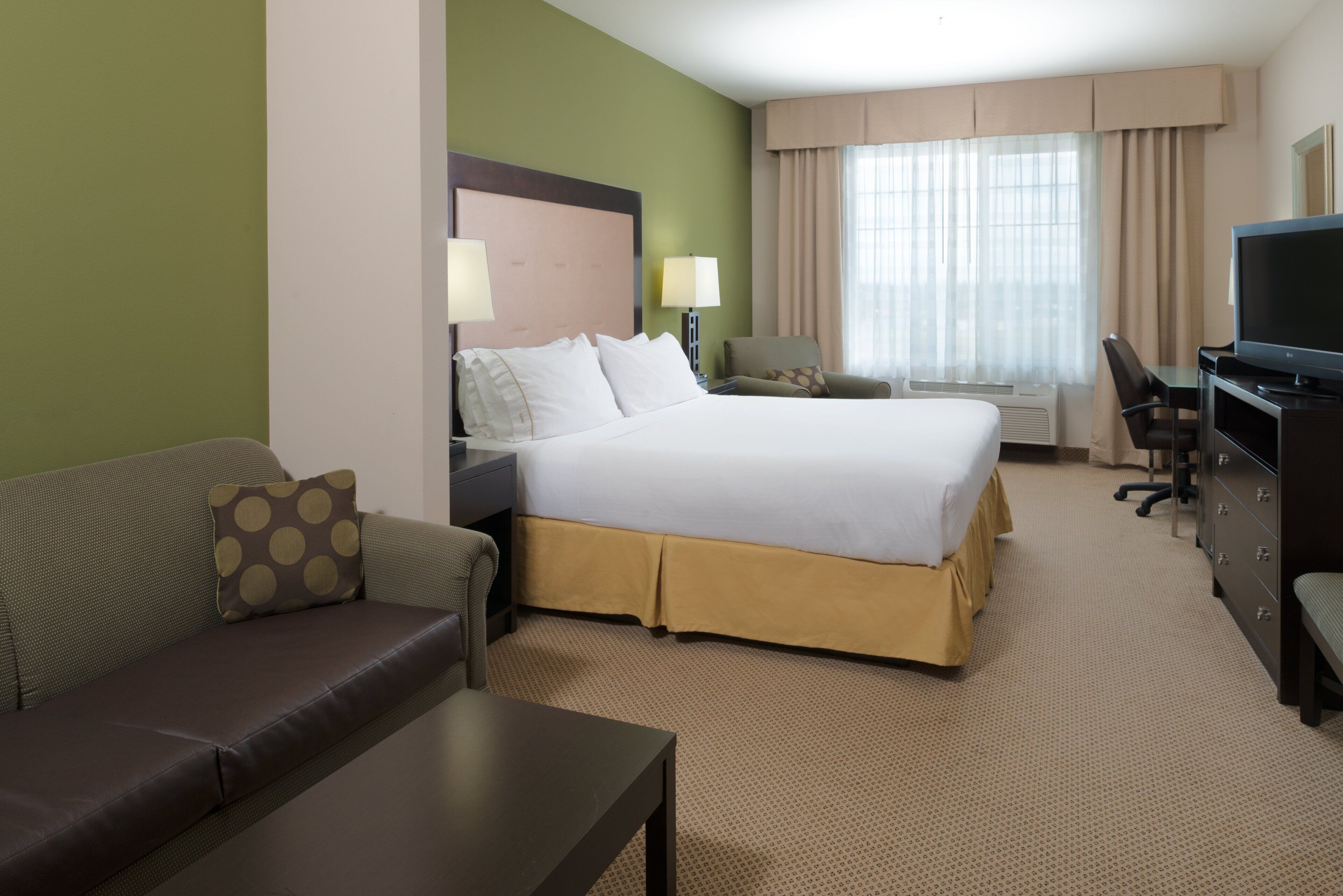 Habitación de hotel con cama, sofá, escritorio, televisión y ventana con cortinas.