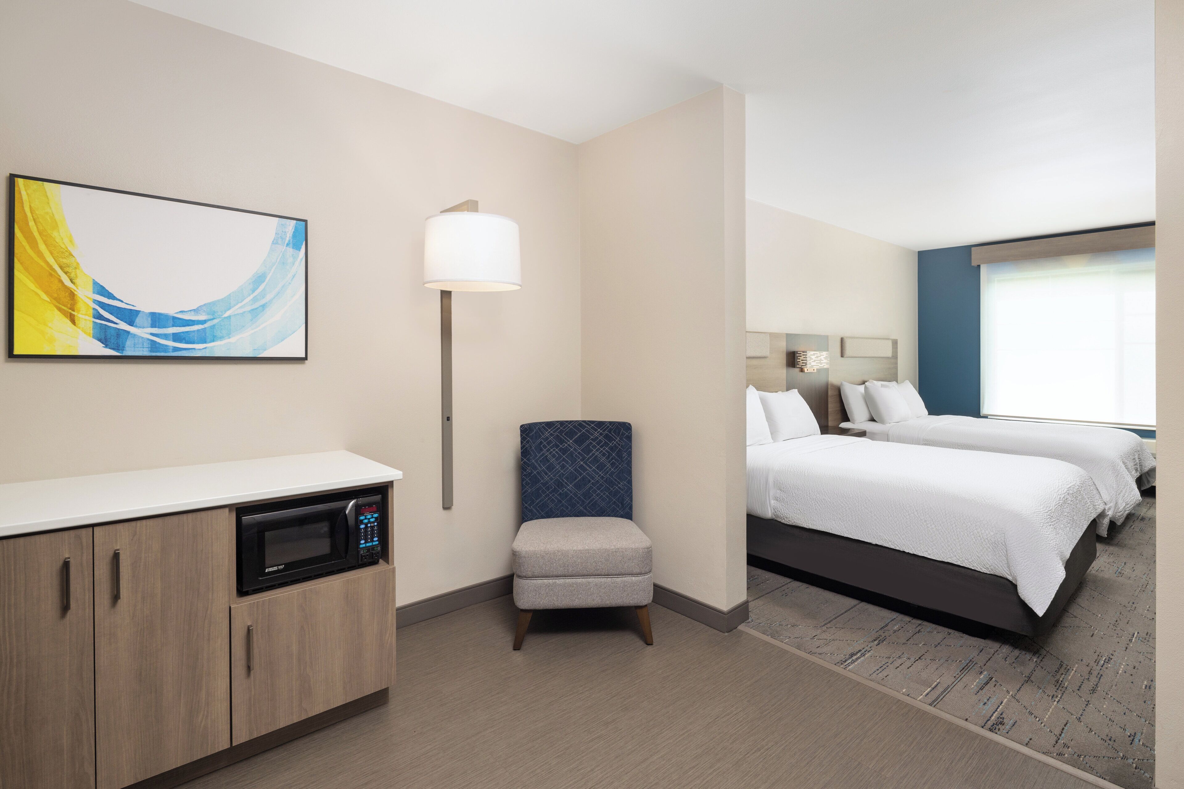 A hotel room with a kitchenette, a single bed, a small armchair, and a large window.