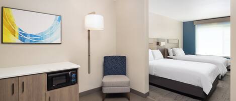 A hotel room with a kitchenette, a single bed, a small armchair, and a large window.