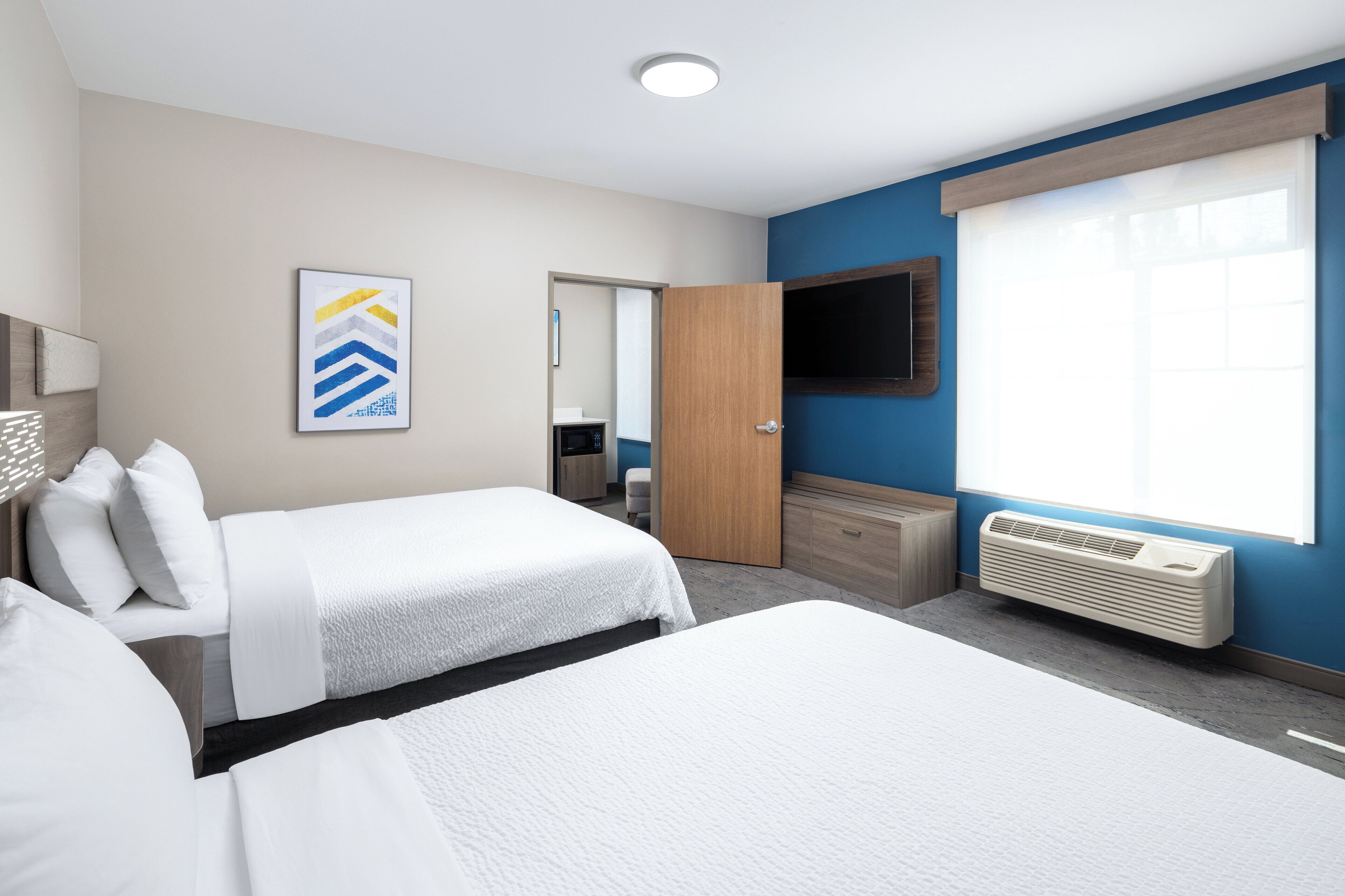 A hotel room with two beds, a flat-screen TV, a large window, and a wall-mounted air conditioner.