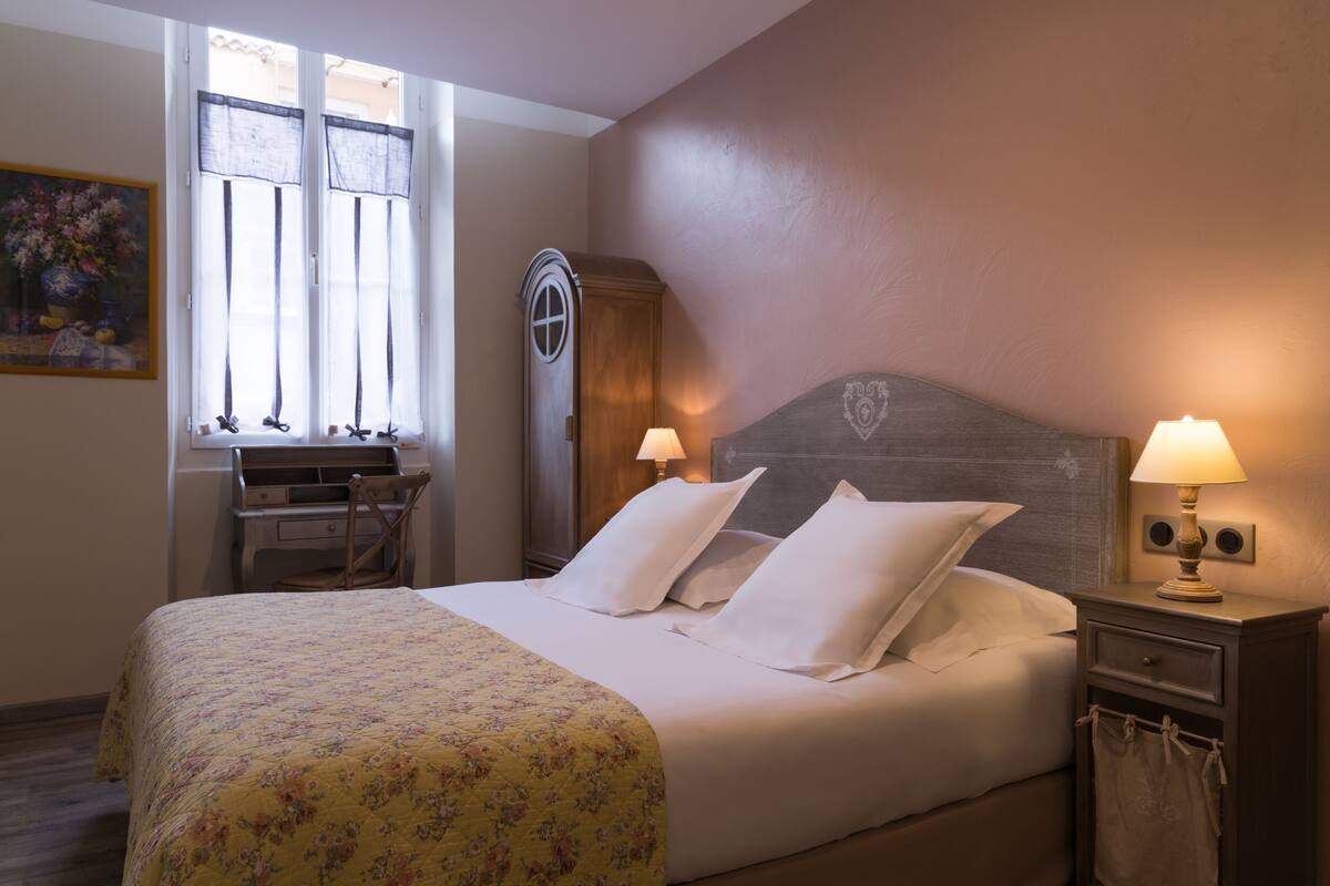 A neatly made bed with white linens and a floral bedspread, a wooden nightstand with a lamp, a wooden wardrobe, and a desk with a chair.