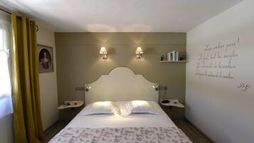 Un dormitorio con cama, dos mesitas de noche, una mesita auxiliar, un estante en la pared y una ventana con cortinas.