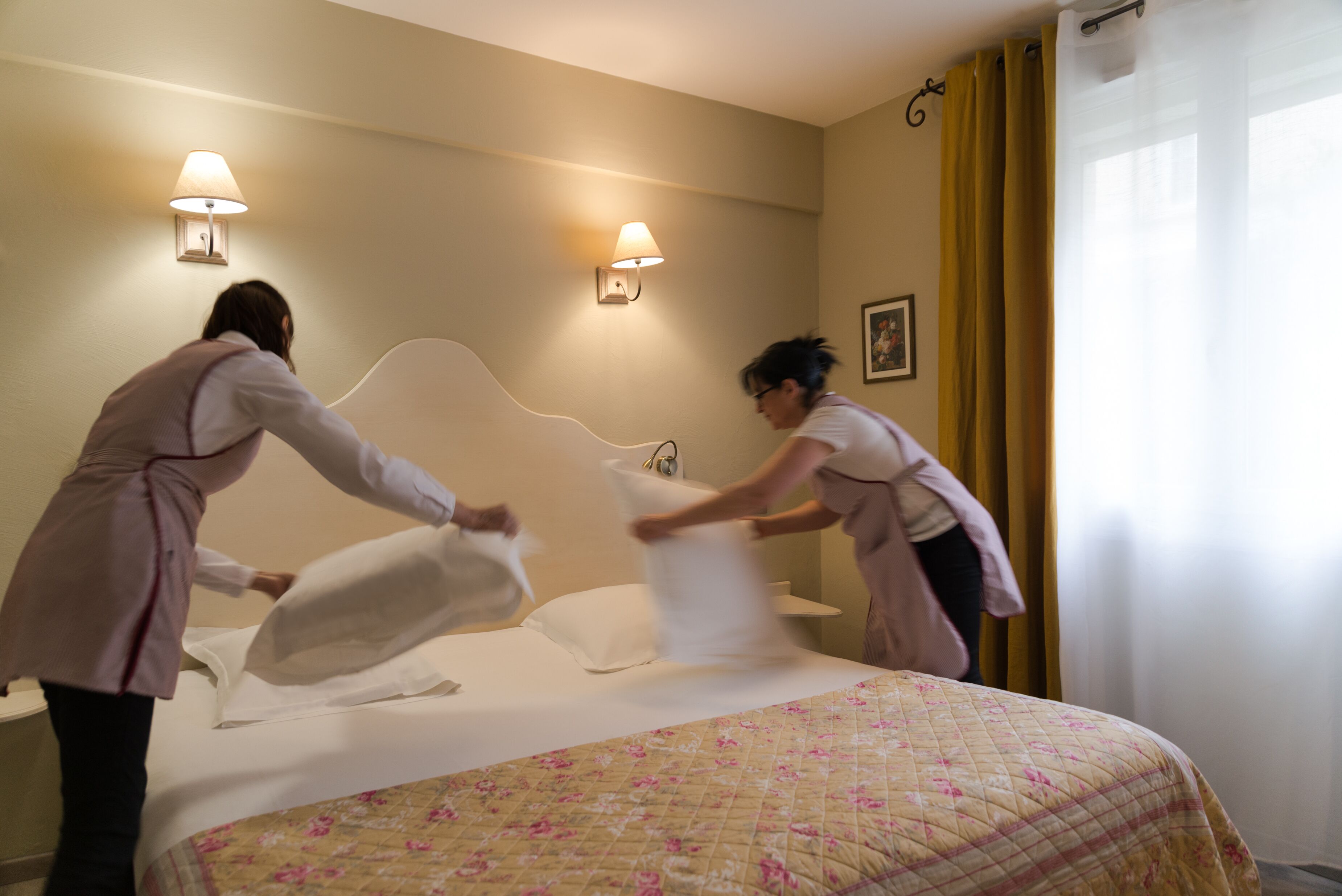 A hotel room with a bed, two wall-mounted lamps, a window with yellow curtains, and a painting on the wall.