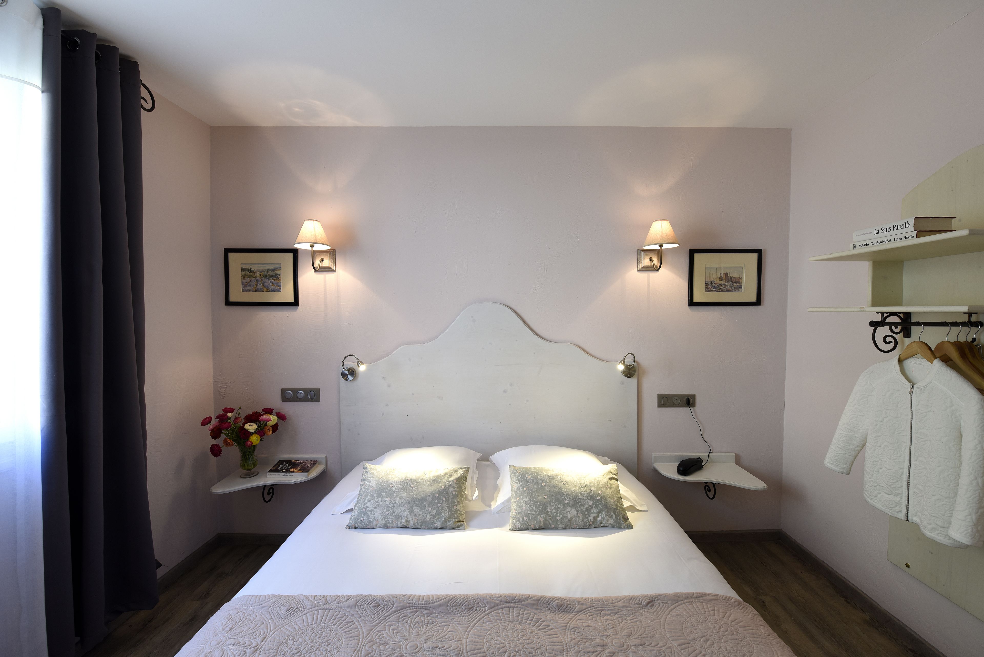 A neatly made bed with two pillows, a bedside table with a lamp, and a shelf with books and a vase of flowers.