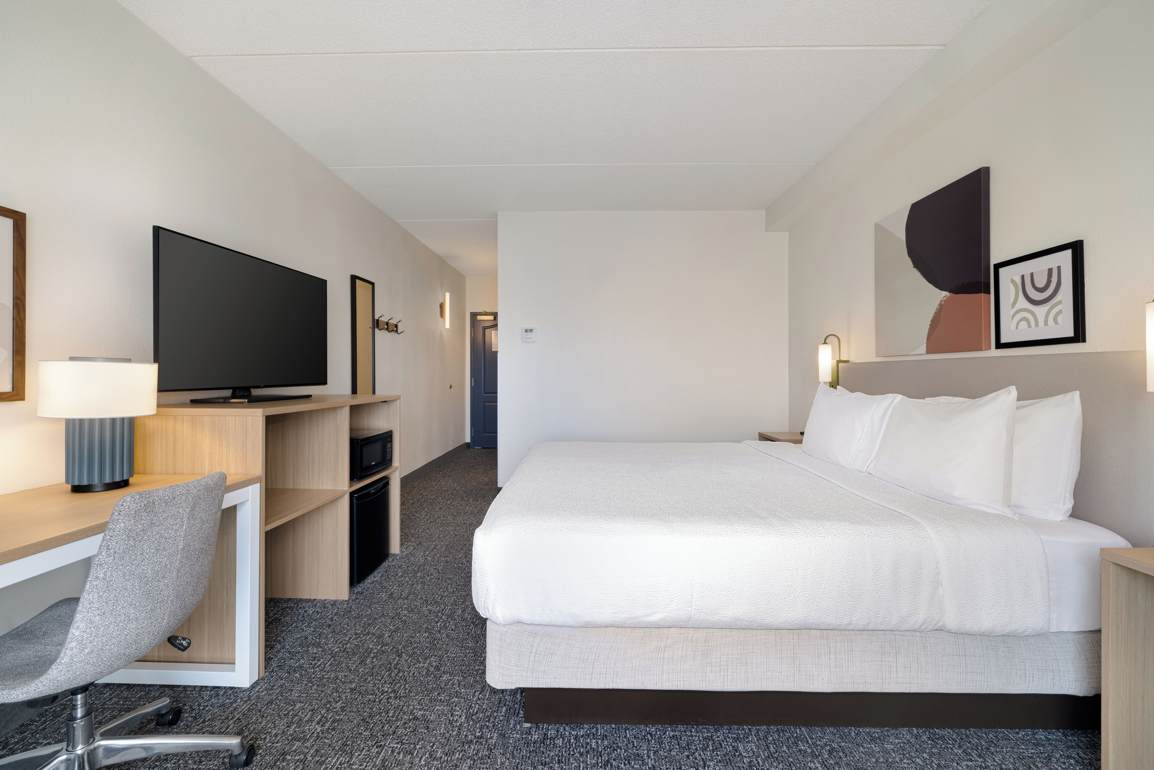 A hotel room with a bed, a desk with a chair, a television, and a wall-mounted light.