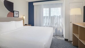 A hotel room with a large bed, a desk, a TV, a window with sheer curtains, and a wall with a framed picture.