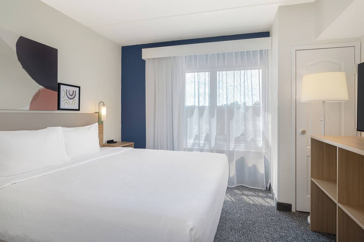 Hotelkamer met een groot bed, een bureau, een televisie, een raam met dunne gordijnen en een wand met een ingelijst kunstwerk.