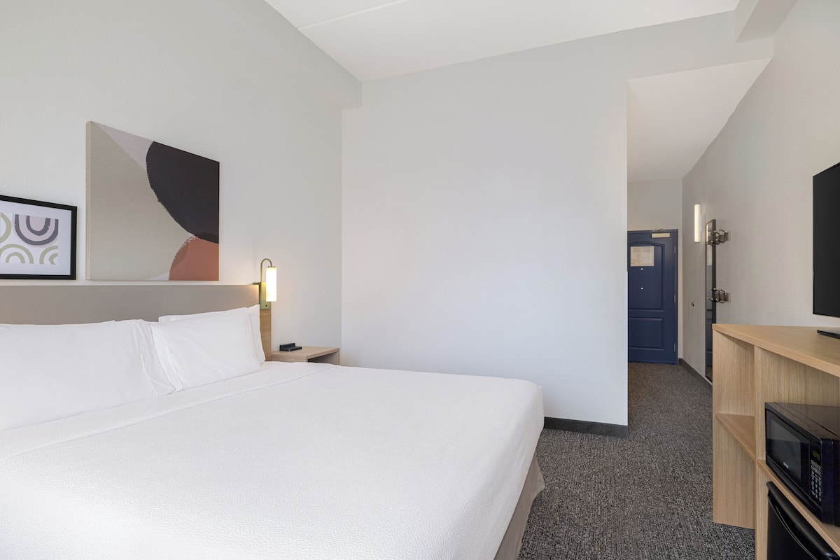 Een hotelkamer met een bed, een nachtkastje, een aan de muur bevestigde televisie en een deur naar een andere kamer.