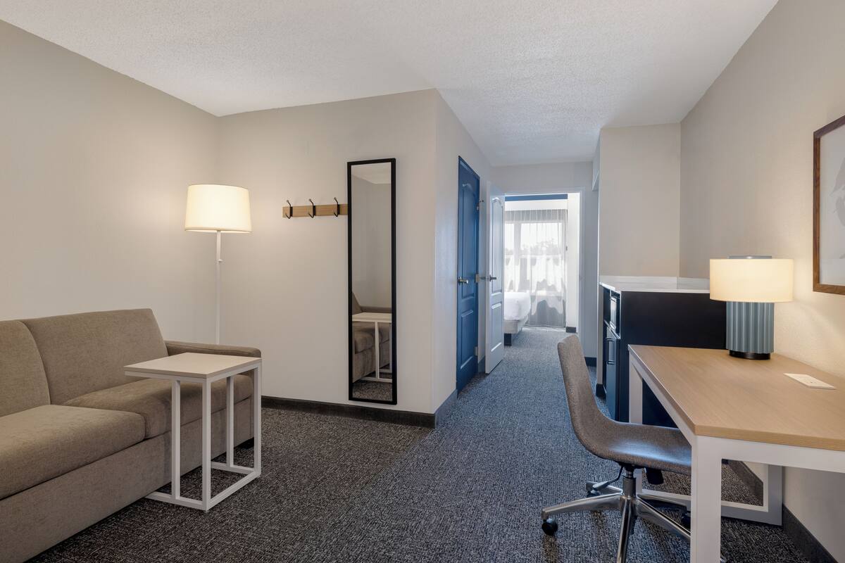 Hotelkamer met een bank, een bureau, een stoel, een lamp, een spiegel en een deur naar een andere kamer.