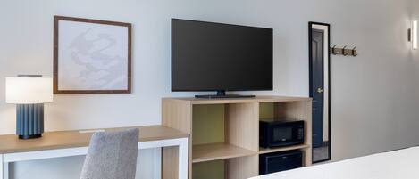 A hotel room with a bed, a desk with a chair, a television, a microwave, and a door with a coat hook.