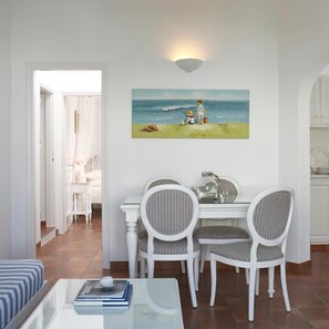 A dining area with a table and chairs, a painting of a beach scene, and a view into a room with a bed and curtains.