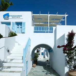 A white building with blue accents, a sign for 'Folegandros apartments,' and a covered outdoor area with a staircase leading to an archway. - Folegandros Apartments (Folegandros)