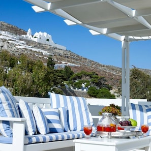 Un área de estar exterior blanca con cojines a rayas, una mesa con una bandeja de frutas, una jarra y vasos, con vistas a una colina con edificios blancos.