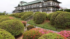 Un edificio tradicional japonés de varias plantas, con una escalera que conduce a la entrada y jardines bien cuidados con arbustos podados y plantas en flor.