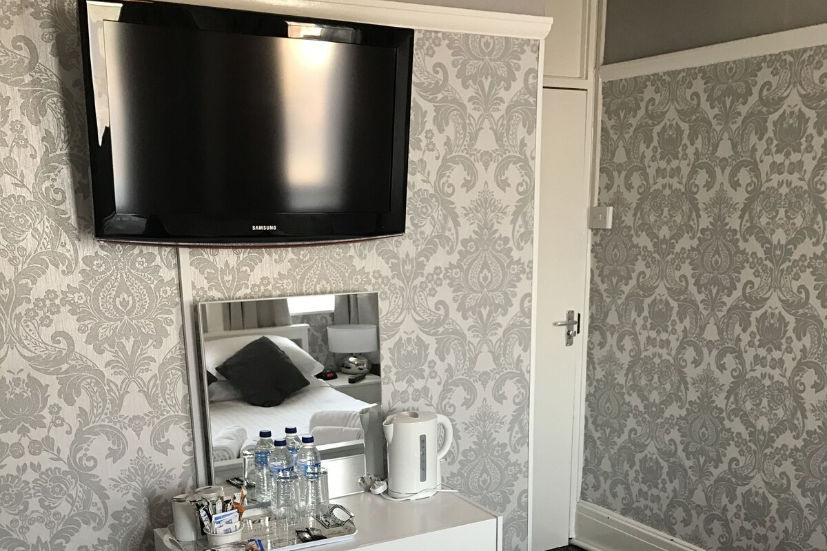 Une chambre d’hôtel avec un lit, un bureau doté d’un miroir, une télévision et une petite poubelle.