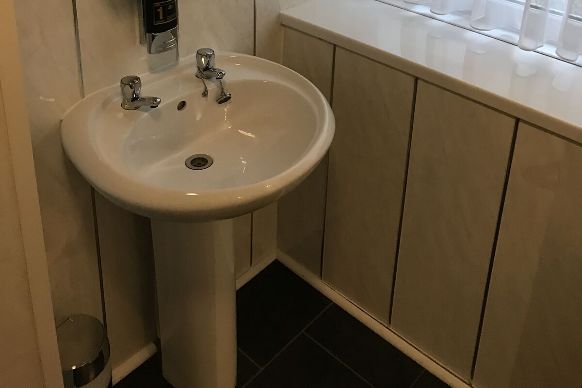 Une salle de bain avec un lavabo blanc, un miroir, un point d’eau et une fenêtre ornée de rideaux légers.