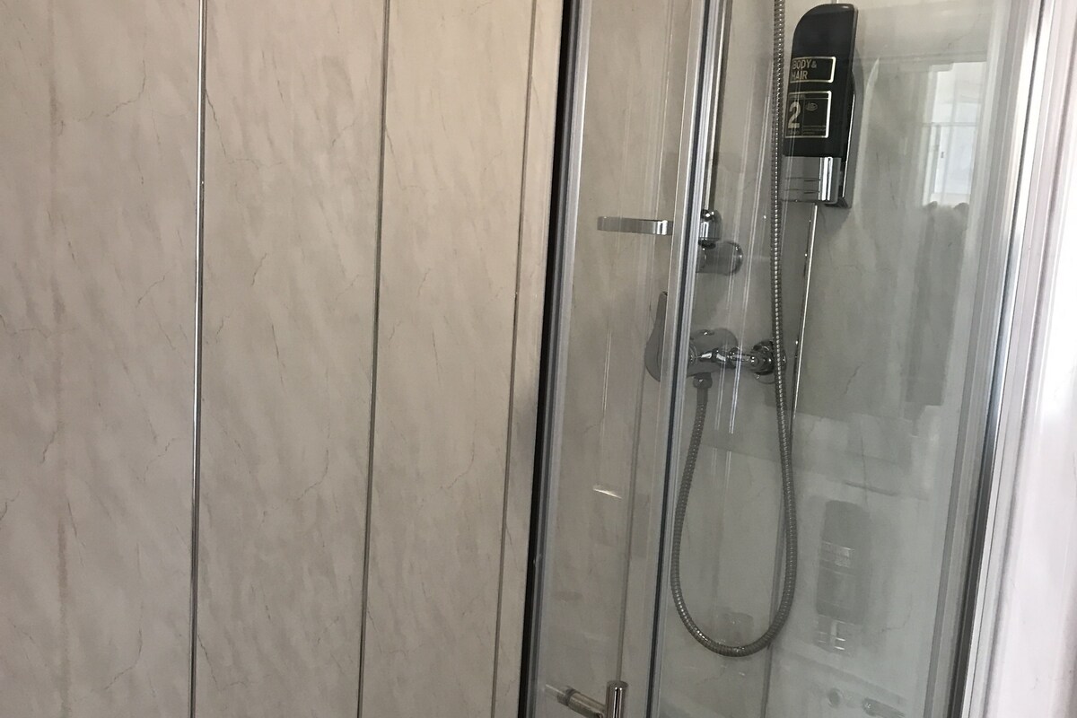 Une douche fermée par des parois en verre, dotée d’un pommeau de douche à main et d’un panneau de commande fixé au mur.
