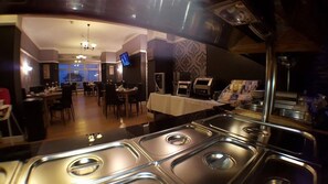 Un espace buffet doté de présentoirs en acier inoxydable, d’un téléviseur fixé au mur et d’un coin repas avec des tables et des chaises.