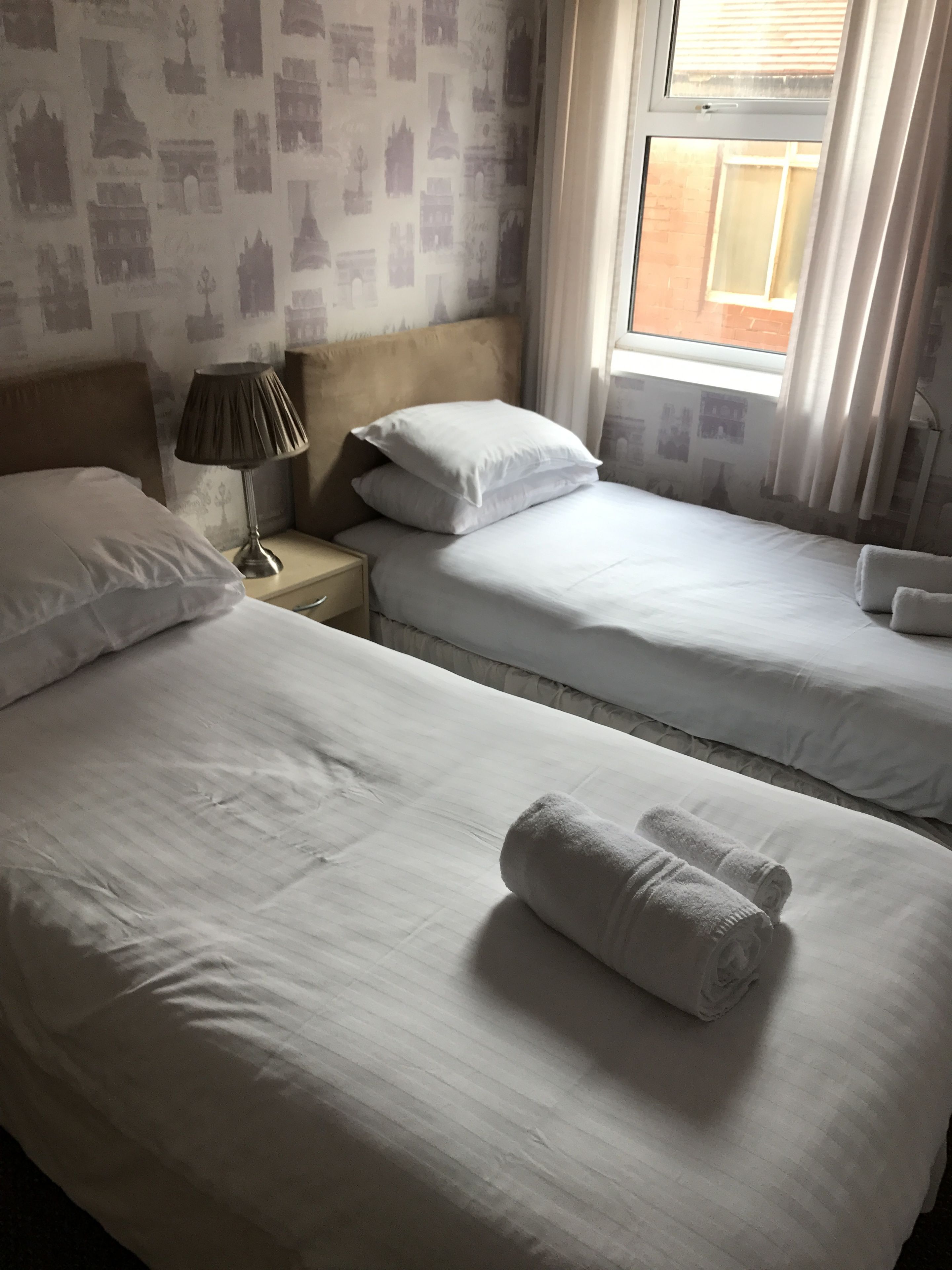 A hotel room with two beds, a nightstand with a lamp, and a window with curtains.