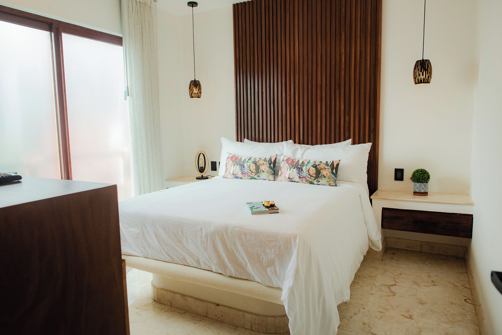 A bedroom with a bed, bedside tables, a nightstand, a small potted plant, and a window with curtains.