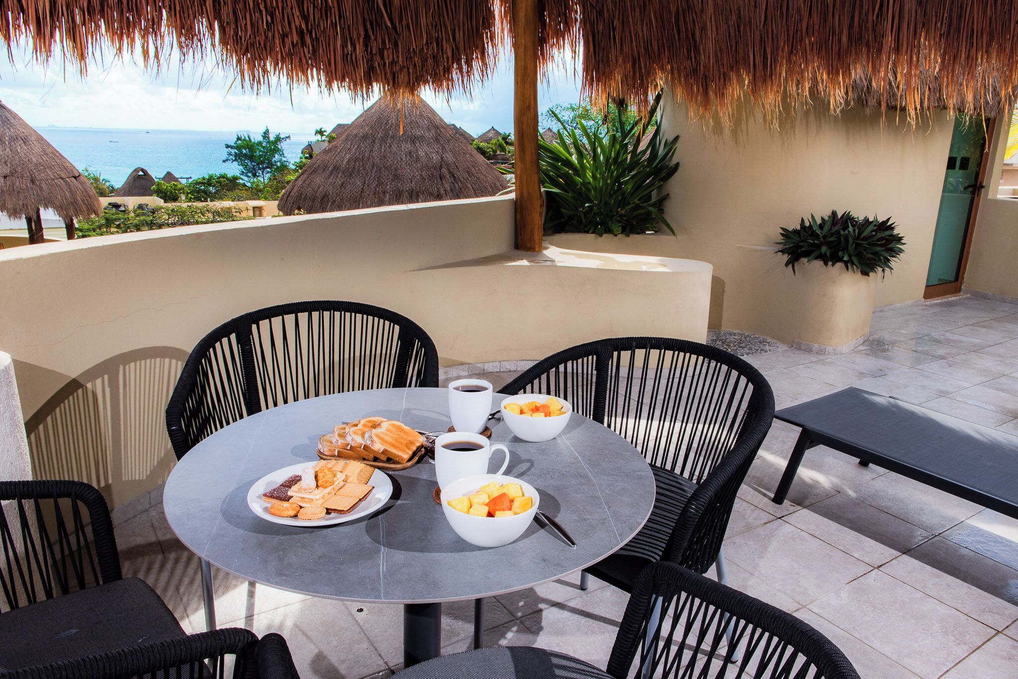 A round table with chairs, a coffee cup, bowls of fruit, and a plate of pastries on a terrace with a view of the sea and thatched-roof buildings.