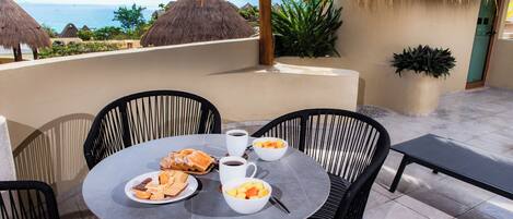 A round table with chairs, a coffee cup, bowls of fruit, and a plate of pastries on a terrace with a view of the sea and thatched-roof buildings.