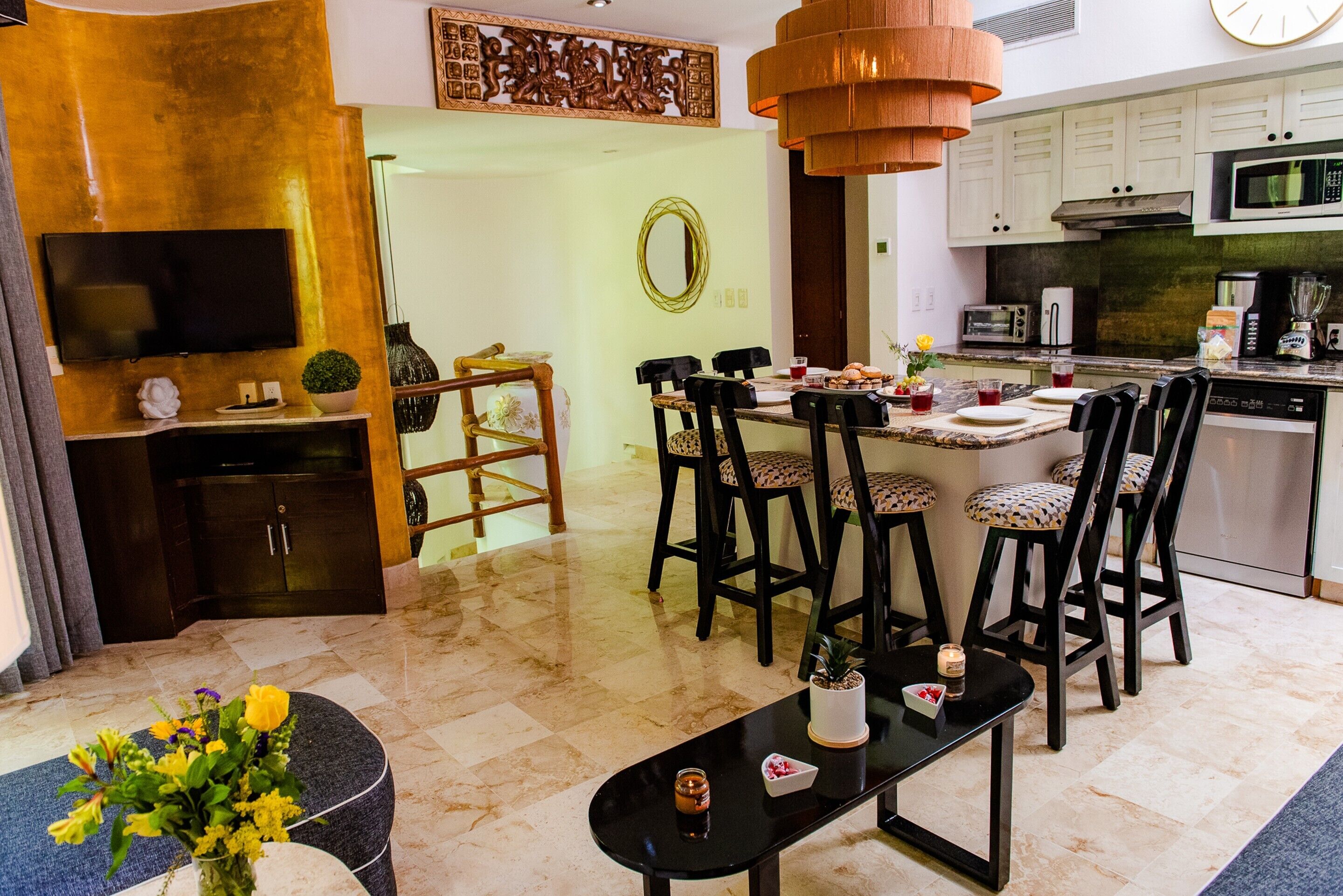 A modern living room with a TV, a dining area with chairs, a kitchen with appliances, and a decorative wall piece.