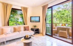 A living room with a sofa, a coffee table, and a TV. There's a balcony with wicker furniture and a view of greenery. - Maya Villa Condo Hotel & Beach Club (Playa del Carmen)