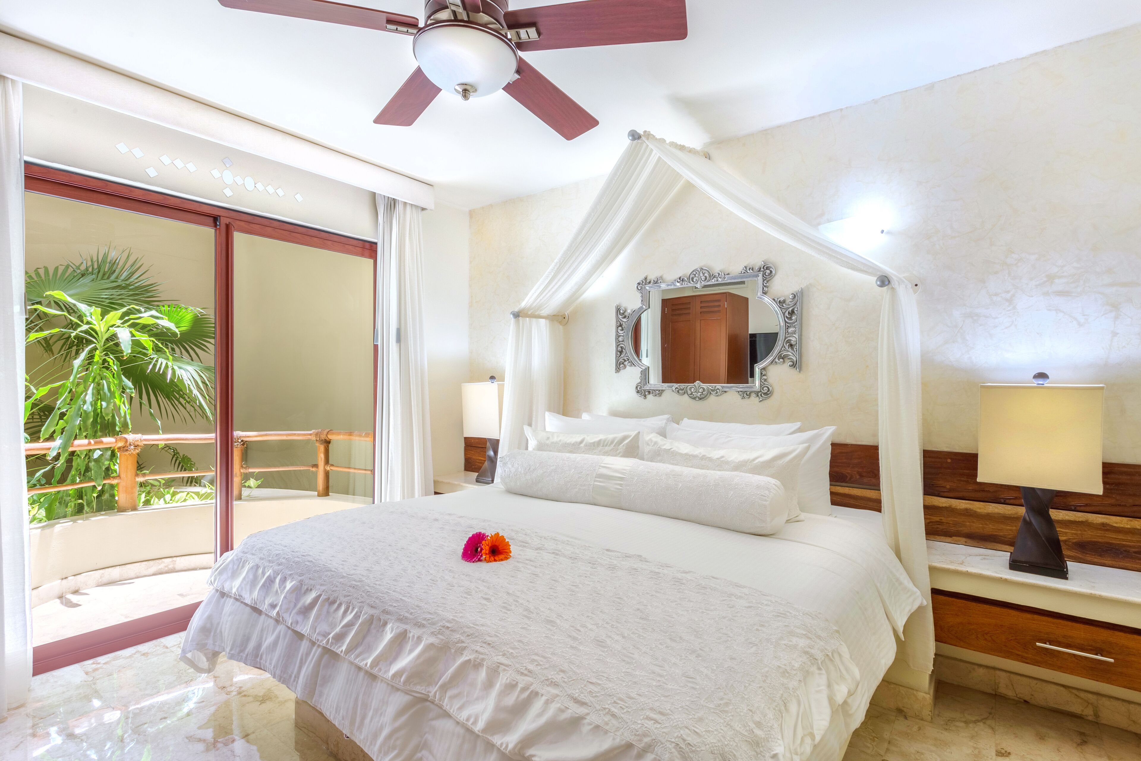 A bedroom with a four-poster bed, a ceiling fan, a large mirror, and a view of a palm tree through a sliding glass door.
