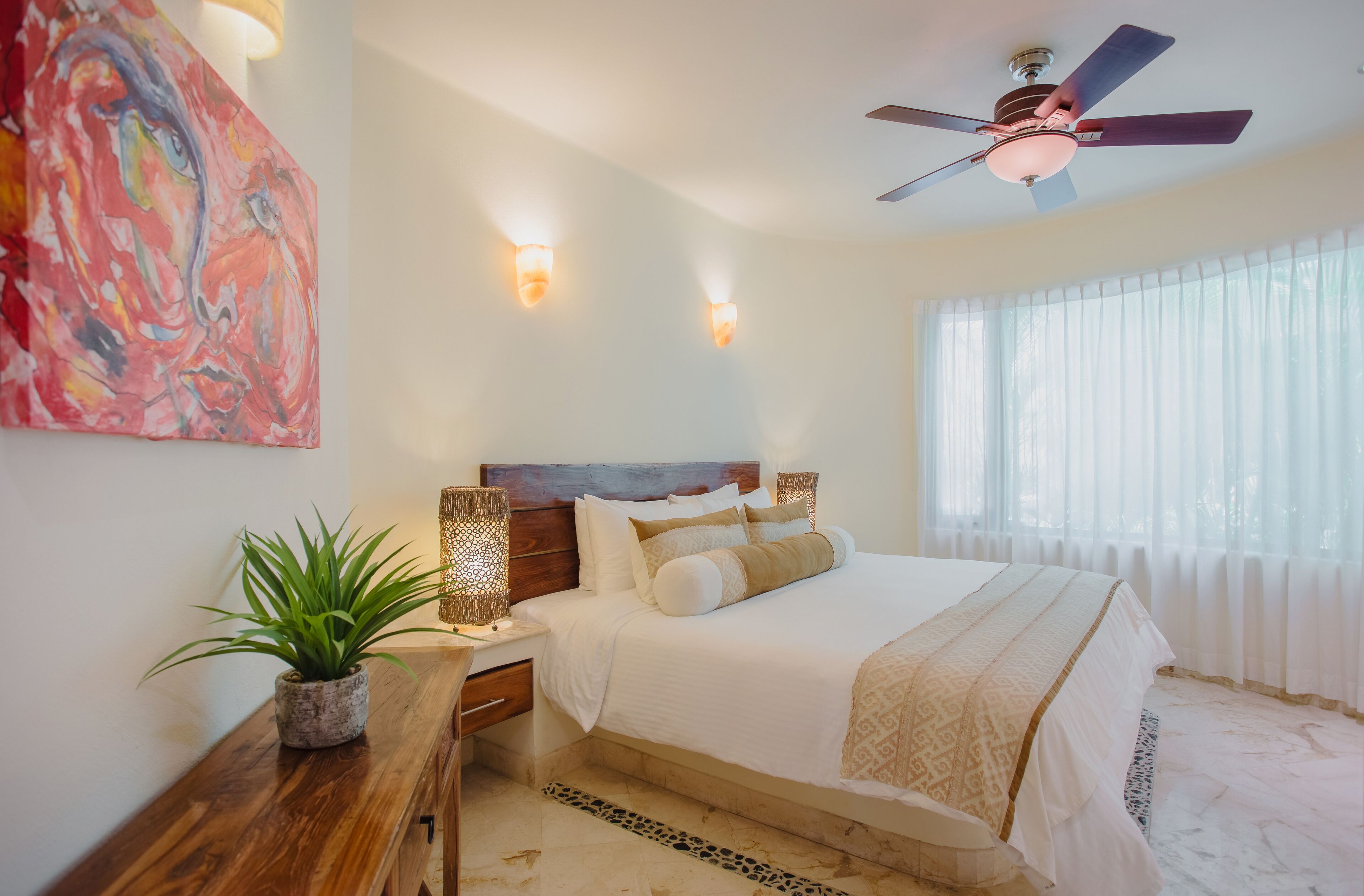 Un dormitorio con cama, mesitas de noche, ventilador de techo y un amplio ventanal con cortinas traslúcidas.