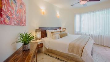 Un dormitorio con cama, mesitas de noche, ventilador de techo y un amplio ventanal con cortinas traslúcidas.
