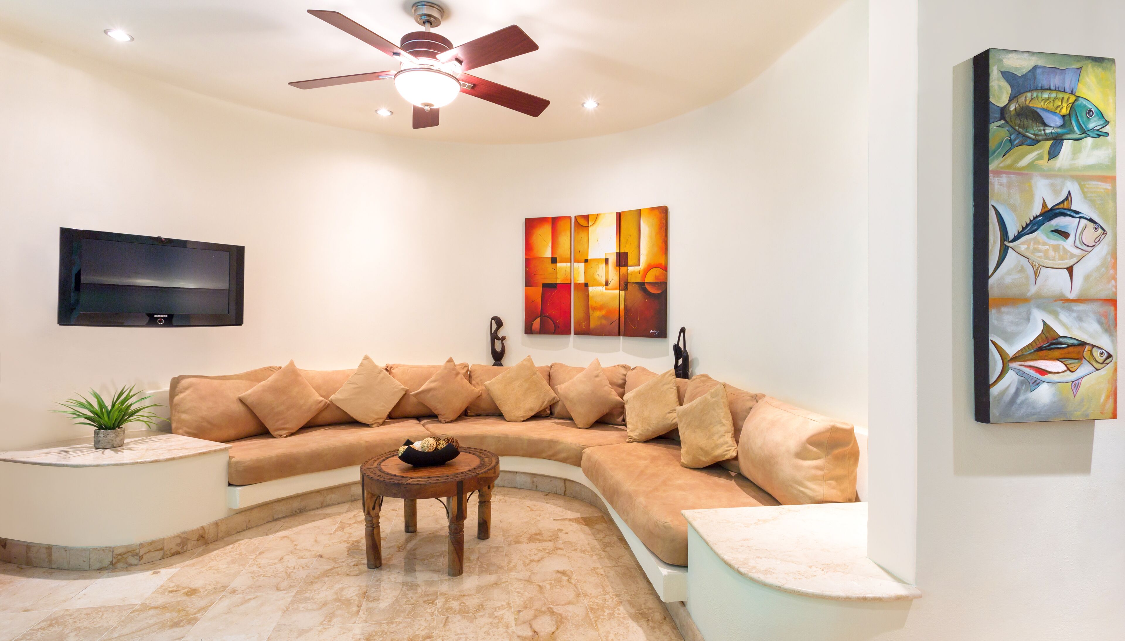 A modern living room with a curved sectional sofa, a ceiling fan, a flat-screen TV, abstract wall art, and a small table with a plant.