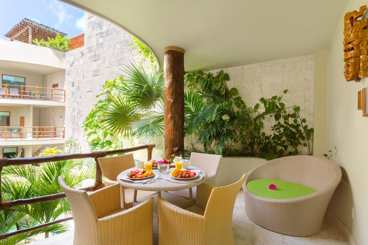 A balcony with wicker furniture, a table set with fruit and drinks, and lush greenery.