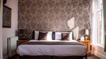 A bedroom with a large bed, floral wallpaper, a radiator, and a window with curtains.