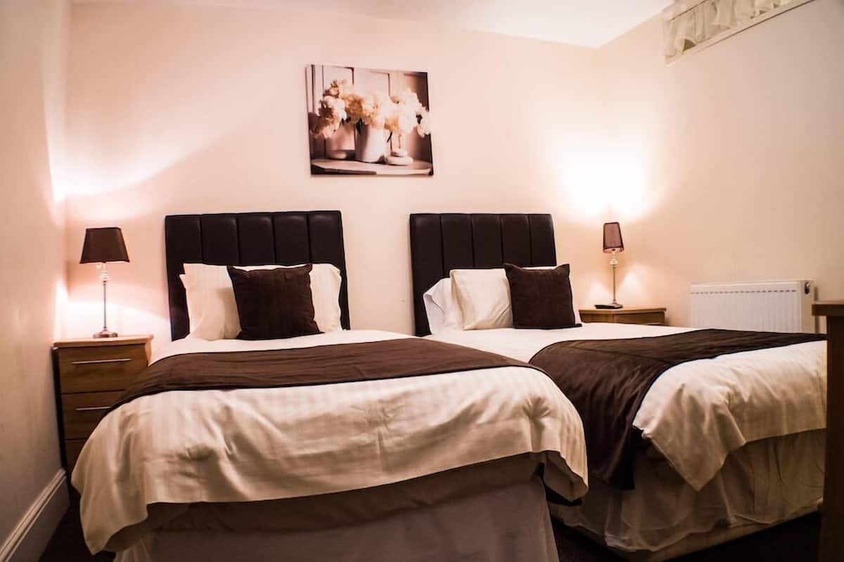 Habitación de hotel con dos camas, cada una con cabecero oscuro y ropa de cama blanca. Hay mesitas de noche con lámparas, un radiador y un cuadro enmarcado en la pared.