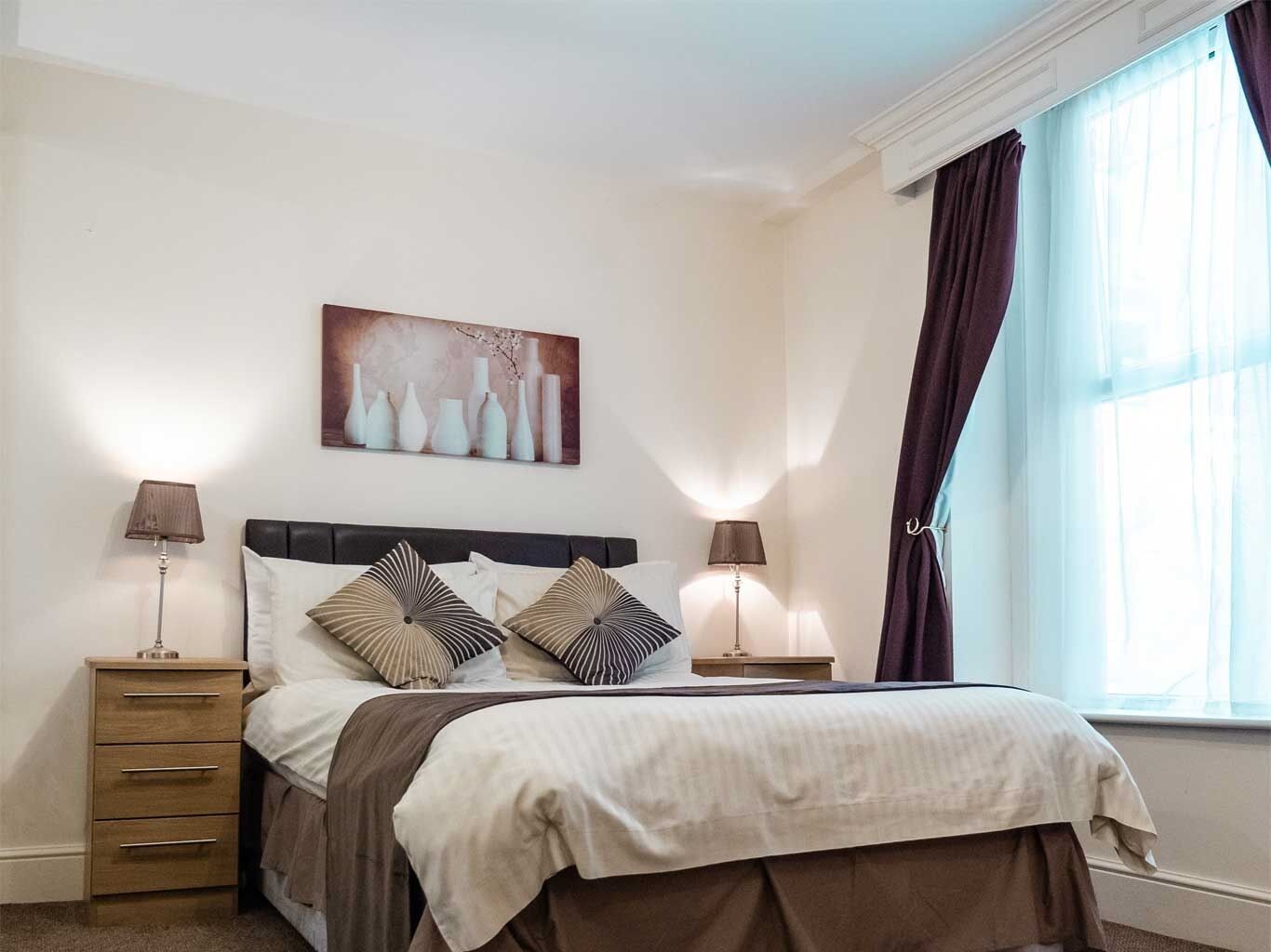 A bedroom with a bed, two bedside tables, a nightstand, a window with curtains, and a wall-mounted artwork.